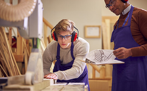 Tutor with male carpentry student in workshop studying for apprenticeship at college using bench saw